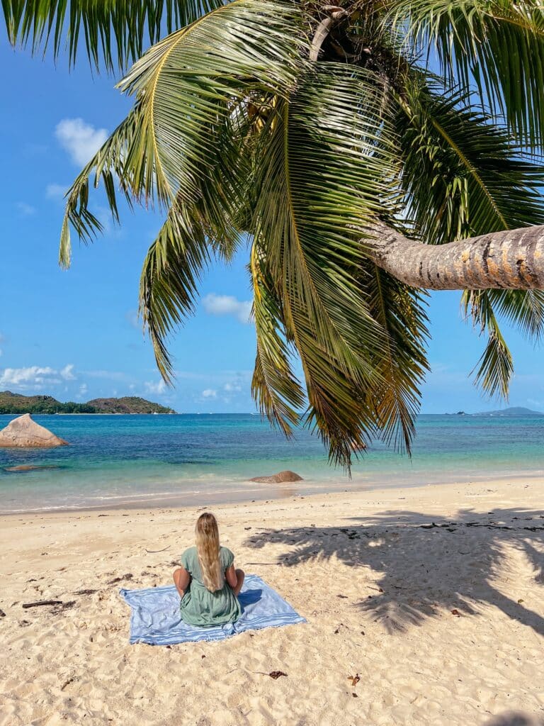 Reisende sitzt unter einer Palme an der Anse Boudin auf Praslin, Seychellen – auf einem Handtuch mit Blick auf das Meer und die Insel Curieuse