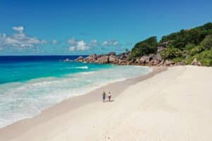 Drohnenaufnahme eines Paares beim Spaziergang an der Grand Anse auf La Digue, Seychellen, mit weißem Sandstrand und türkisfarbenem Meer