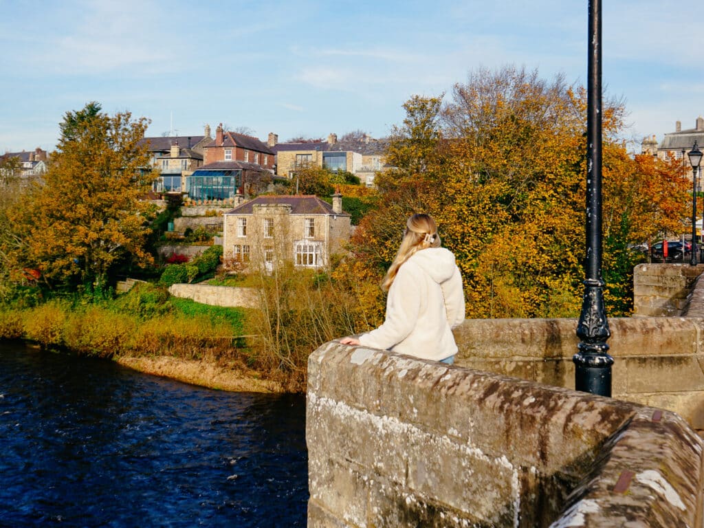 Frau steht auf der historischen Steinbrücke von Corbridge in Northumberland mit Blick auf das Dorf und herbstliche Landschaft